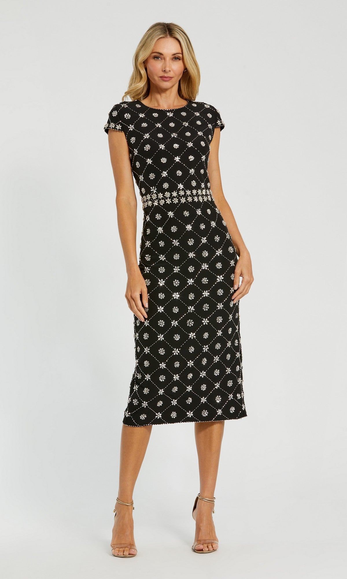 A woman wears the Mac Duggal 2234 black short-sleeve knee-length dress with a white geometric pattern—an elegant Mauve Pink Wedding Guest Dress option—styled with nude high-heeled sandals against a plain white background.